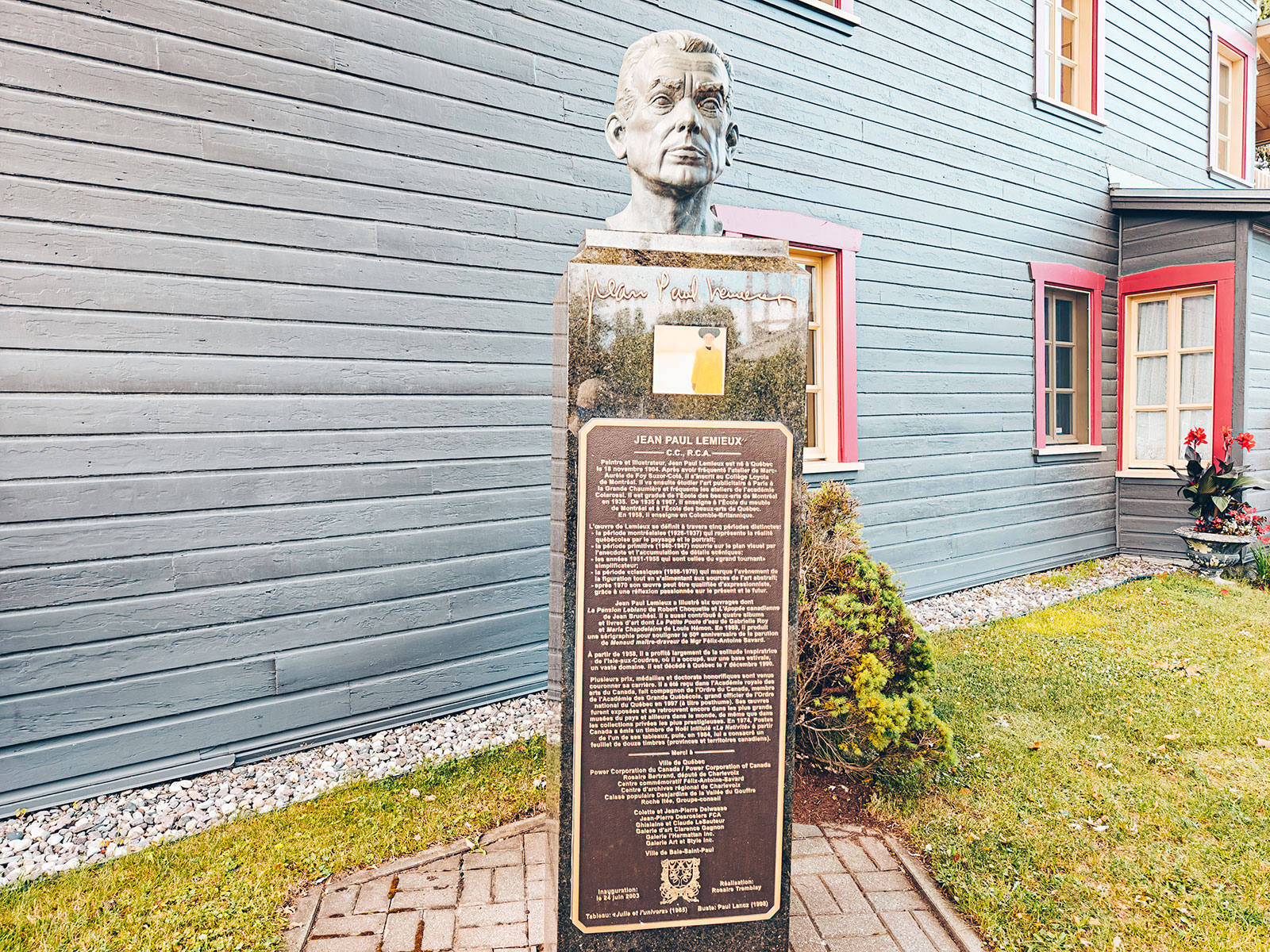 Un buste sur socle avec sa plaque, devant un bâtiment bleu
