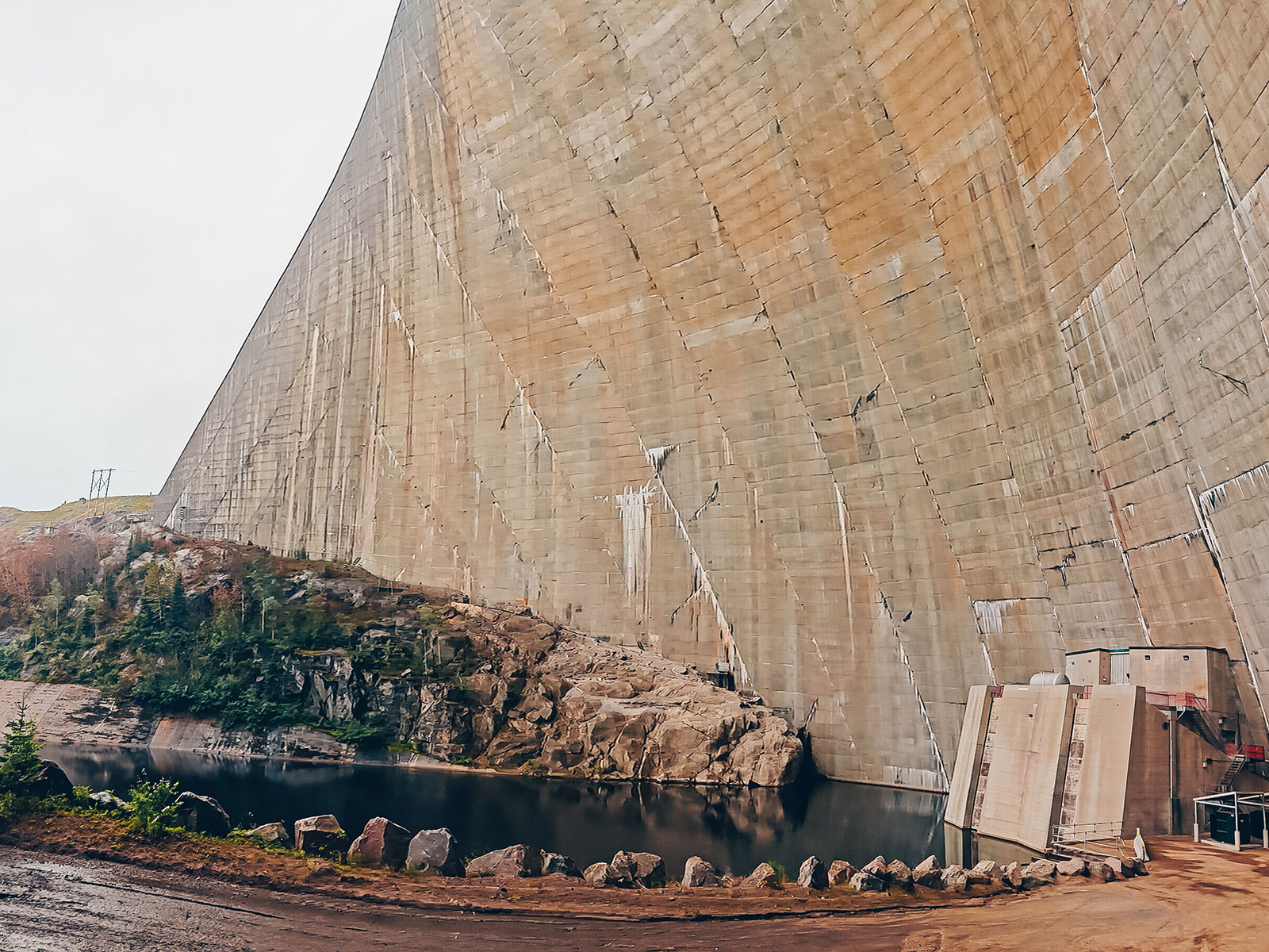 Contre-plongée sur le parement de béton incurvé du barrage, un petit bâtiment technique à sa base