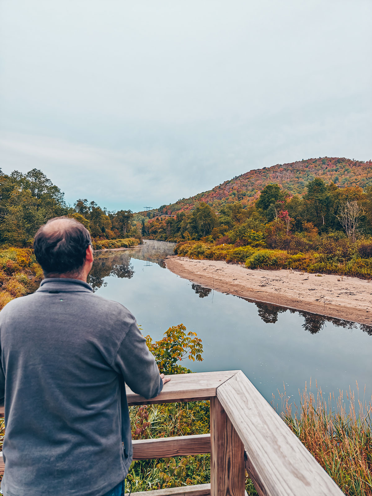 Un homme de dos, accoudé à la rambarde, regarde la rivière et les collines